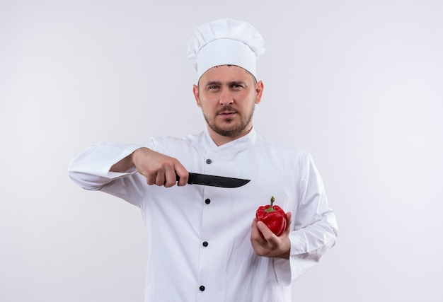 Confident young handsome cook in chef uniform holding pepper and pointing with knife at it isolated on white wall