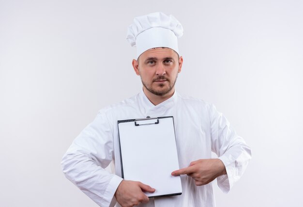 Confident young handsome cook in chef uniform holding and pointing at clipboard isolated on white wall