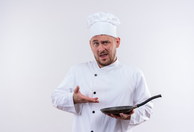 Confident young handsome cook in chef uniform holding and pointing at frying pan isolated on white wall