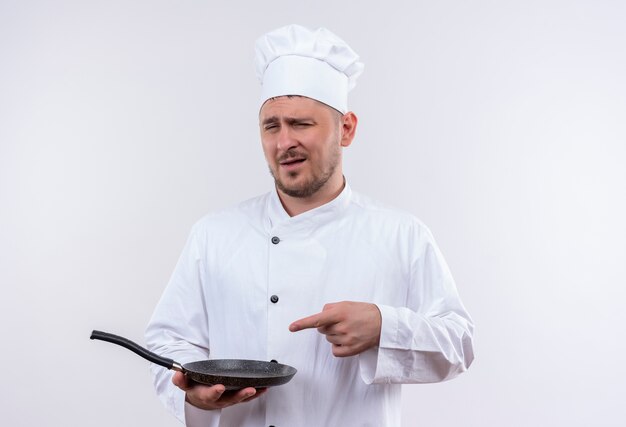 Confident young handsome cook in chef uniform holding and pointing at frying pan isolated on white wall