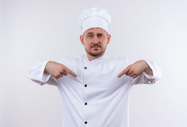 Confident young handsome cook in chef uniform pointing at himself isolated on white wall