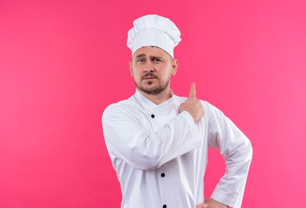 Уверенный молодой красивый повар в униформе шеф-повара, указывая позади изолированной на розовой стене