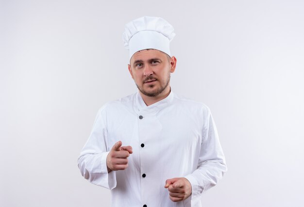 Confident young handsome cook in chef uniform pointing isolated on white wall