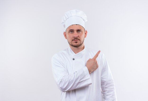 Confident young handsome cook in chef uniform pointing at side isolated on white wall