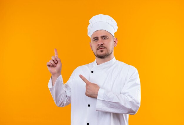 Confident young handsome cook in chef uniform pointing up isolated on orange wall