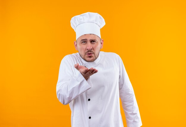 Confident young handsome cook in chef uniform sending blow kiss isolated on orange wall