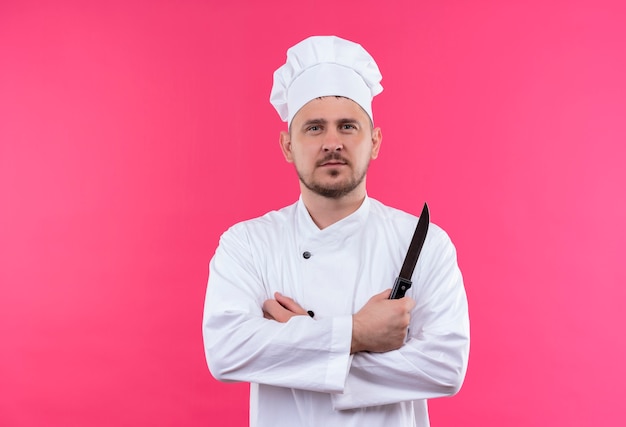 閉じた姿勢で立って、ピンクの壁にナイフを保持しているシェフの制服を着た自信のある若いハンサムな料理人