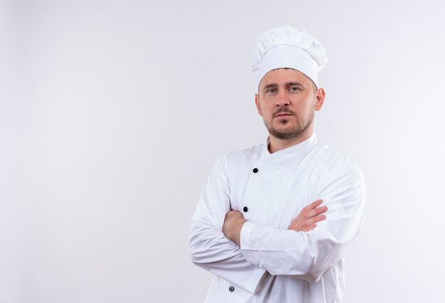 Confident young handsome cook in chef uniform standing with closed posture isolated on white wall