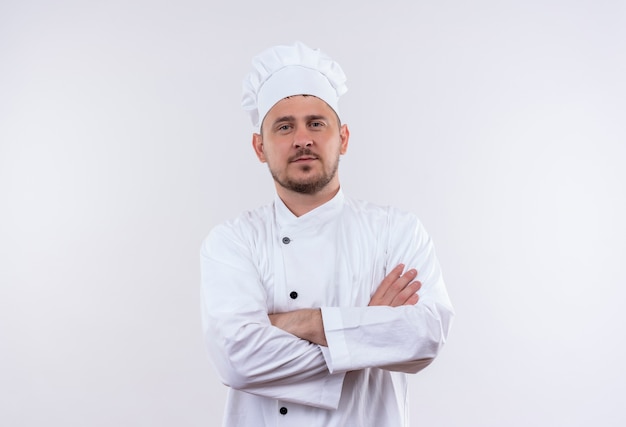 Confident young handsome cook in chef uniform standing with closed posture isolated on white wall
