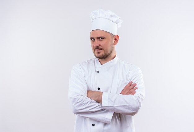 Confident young handsome cook in chef uniform standing with closed posture isolated on white wall