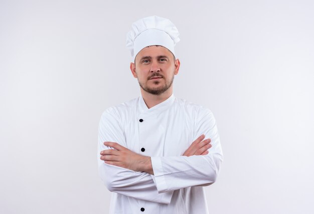 Confident young handsome cook in chef uniform standing with closed posture isolated on white wall