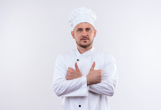 Confident young handsome cook in chef uniform standing with closed posture showing thumbs up isolated on white wall