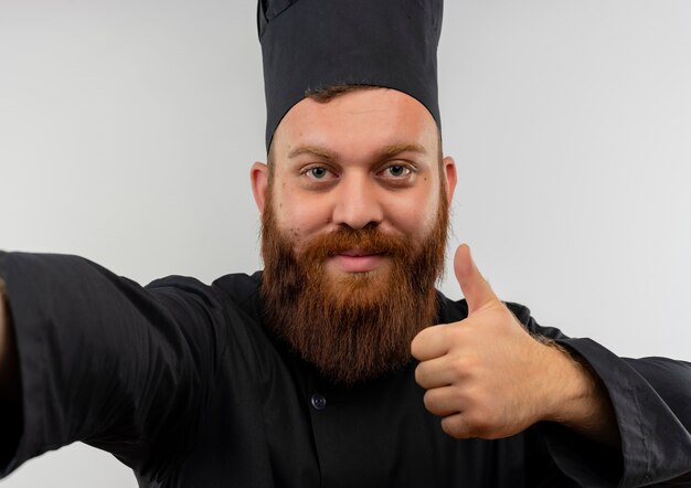 Confident young handsome cook in chef uniform stretching out hand and showing thumb up isolated on white wall