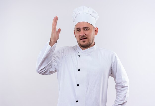Уверенный молодой красивый повар в униформе шеф-повара с поднятой рукой, изолированной на белой стене