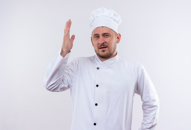 Confident young handsome cook in chef uniform with raised hand isolated on white wall