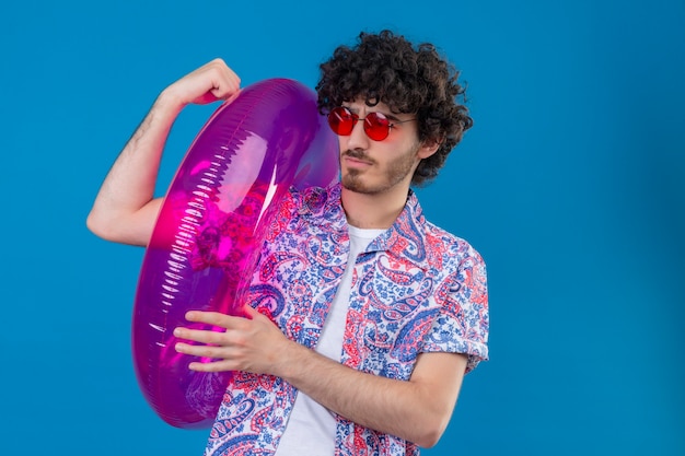 Confident young handsome curly man wearing sunglasses holding swim ring doing strong gesture on isolated blue space with copy space