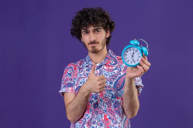 Confident young handsome curly traveler man holding alarm clock showing thumb up on isolated purple space with copy space