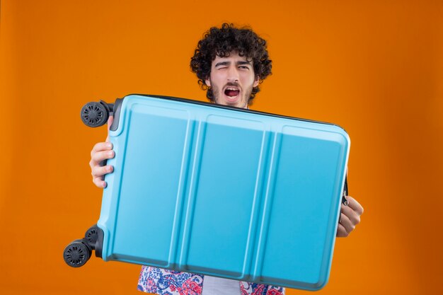 Confident young handsome curly traveler man holding suitcase and winking with open mouth on isolated orange space