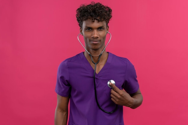 심장 박동을 확인하기 위해 청진기를 사용하여 보라색 유니폼을 입고 곱슬 머리를 가진 자신감이 젊은 잘 생긴 어두운 피부 의사
