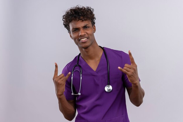 Free photo a confident young handsome dark-skinned doctor with curly hair wearing violet uniform with stethoscope holding hands in the rock gesture