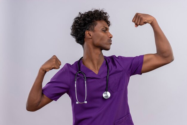 強さのジェスチャーを示す聴診器で紫の制服を着た巻き毛の自信を持って若いハンサムな浅黒い医者