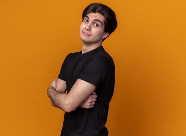 Confident young handsome guy wearing black t-shirt crossing hands isolated on orange wall with copy space