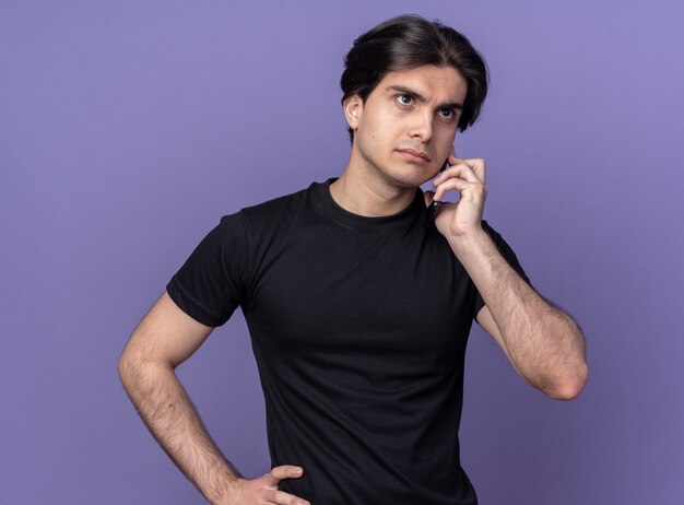 Confident young handsome guy wearing black t-shirt speaks on phone isolated on purple wall