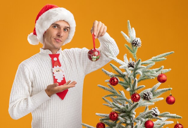 confident young handsome guy wearing christmas hat and santa claus tie standing near decorated christmas tree holding and pointing with hand at christmas ball ornament looking  isolated on orange wall