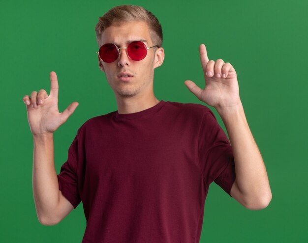 Confident young handsome guy wearing red shirt and glasses points at up isolated on green wall