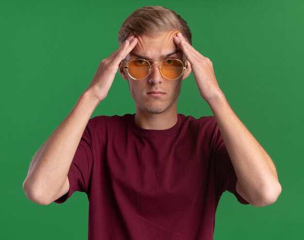 Confident young handsome guy wearing red shirt and glasses putting hands on forehead isolated on green wall