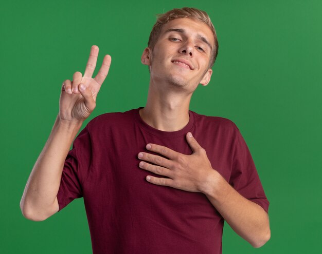 Confident young handsome guy wearing red shirt showing peace gesture putting hand on heart isolated on green wall