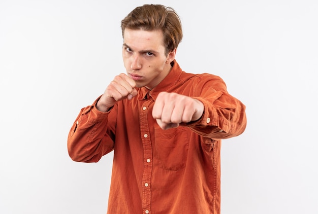 Confident young handsome guy wearing red shirt standing in fighting pose isolated on white wall