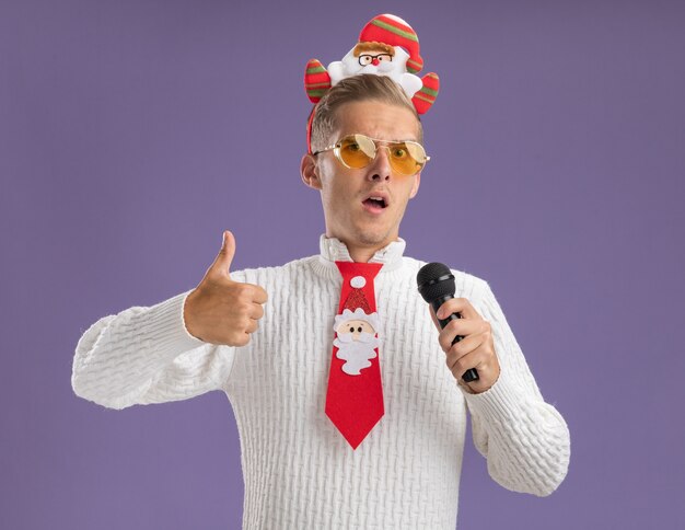 Confident young handsome guy wearing santa claus headband and tie with glasses holding microphone looking at camera showing thumb up isolated on purple background