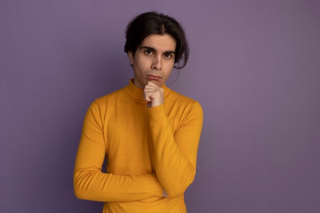 Confident young handsome guy wearing yellow turtleneck sweater grabbed chin isolated on purple wall with copy space