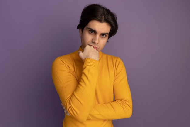 Fiducioso giovane bel ragazzo che indossa maglione dolcevita giallo che mette il pugno sul mento isolato sulla parete viola