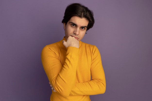 Уверенный молодой красивый парень в желтом свитере с высоким воротом, положив кулак на подбородок, изолированный на фиолетовой стене