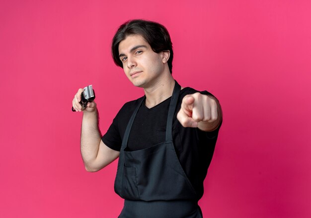 Confident young handsome male barber in uniform holding hair clippers and showing you gesture isolated on pink wall