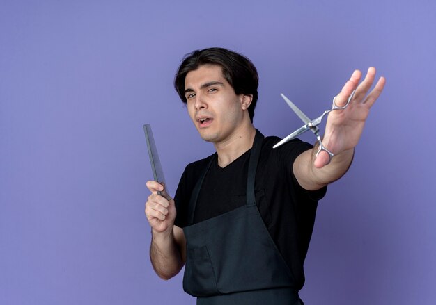 Confident young handsome male barber in uniform holding out scissors at camera  and comb isolated on blue background with copy space