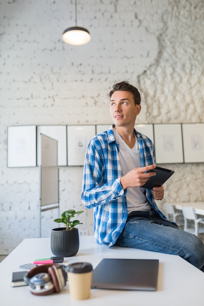 Confident young handsome man in chekered shirt sitting on table using tablet computer in co-working office