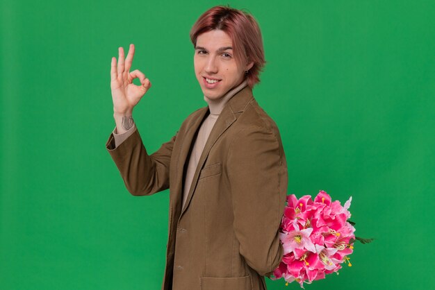 Confident young handsome man holding bouquet of flowers behind his back and gesturing ok sign 