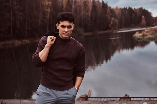 A confident young handsome man holds his coat on a shoulder near a lake in the autumn forest.