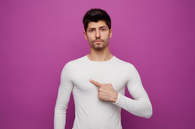 Confident young handsome man looking at camera pointing at himself on purple background