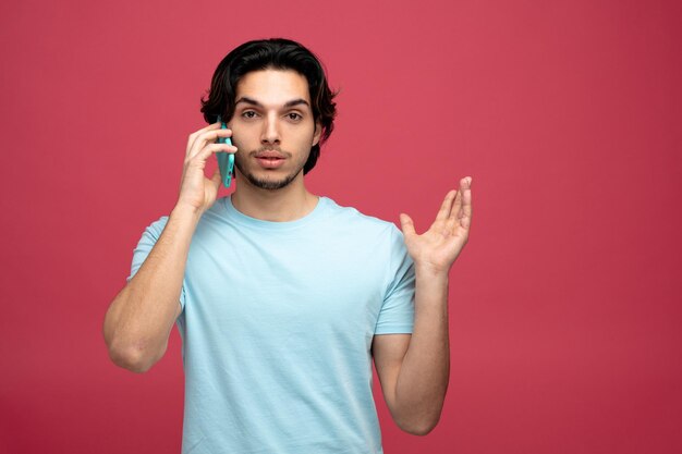 confident young handsome man looking at camera showing empty hand while talking on phone isolated on red background with copy space