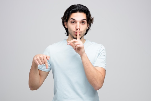 Confident young handsome man looking at camera showing silence gesture pointing down isolated on white background