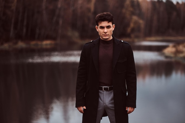 A confident young handsome man standing near a lake in the autumn forest.