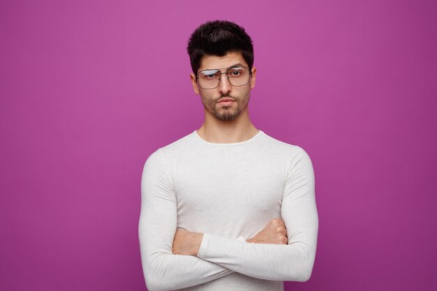 Confident young handsome man wearing glasses looking at camera while keeping arms crossed on purple background