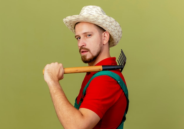 Confident young handsome slavic gardener in uniform and hat standing in profile view holding rake on shoulder looking isolated