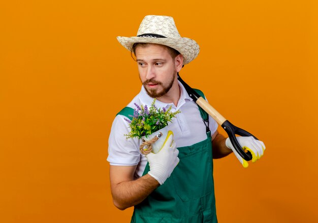 Confident young handsome slavic gardener in uniform wearing hat and gardening gloves holding spade on shoulder and holding flowerpot looking at side isolated