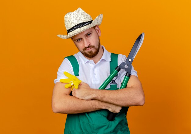 Confident young handsome slavic gardener in uniform wearing hat standing with closed posture holding gardening gloves and pruners looking isolated