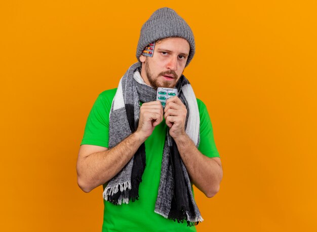 Уверенный молодой красивый славянский больной мужчина в зимней шапке и шарфе показывает пачку капсул с другой пачкой капсул под шляпой, изолированной на оранжевой стене с копией пространства